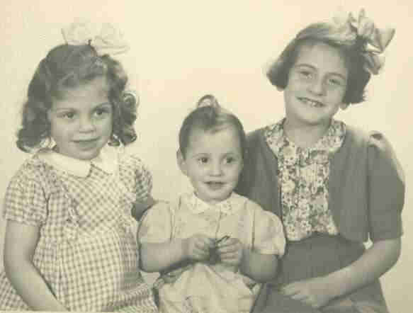 Three young girls sitting in a line - the youngest is in the middle. They are all nicely dressed and smiling. Two older girls insides have bows in their hair.