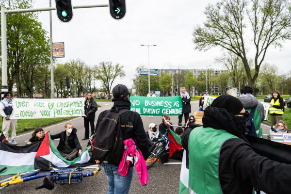 A group of about 15 protests blocks a highway exit by sitting and standing in the middle of the road. They hold Palestinian flags and banners with texts including (in English and Dutch): 'Being stuck at a checkpoint is a daily reality for Palestinian people', and 'No business as usual during genocide'