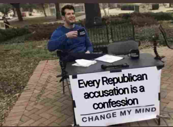 Man holding a cup while sitting in a park at a table which bears a sign reading "Every Republican accusation is a confession. CHANGE MY MIND."