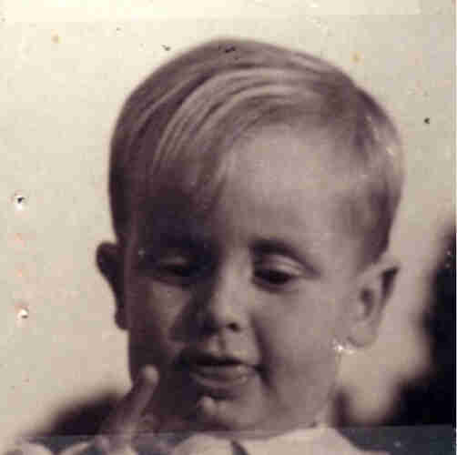Headshot of a young boy. His blond hair is combed over his forehead. He is looking at his fingers. His tongue is slightly extended. 