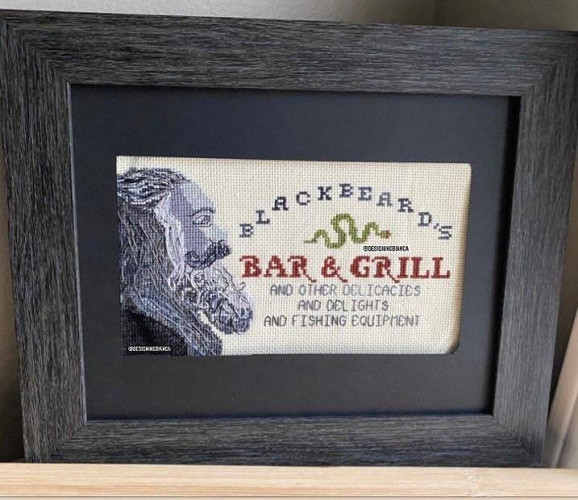 Still image. Framed cross-stitch of a person with long salt-and-pepper hair, full mustache and beard, in profile. The text design beside them reads:
BLACKBEARD'S
[snake graphic]
BAR & GRILL
[in smaller letters]
and other delicacies
and delights
and fishing equipment