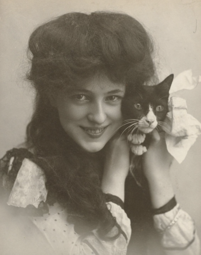 Black and white photo of a beautiful young white woman with voluminous, long dark hair, smiling at the camera and snuggling a shorthaired black tuxedo cat up to the side of her face.