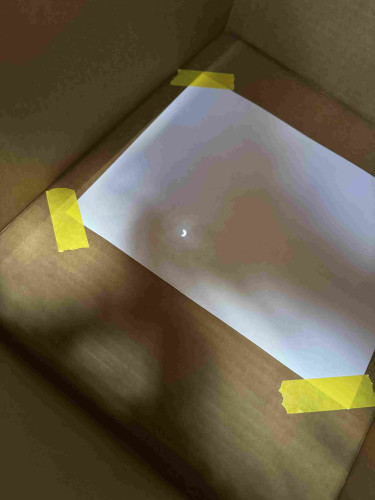 A photo from inside a makeshift camera obscura made from a cardboard box, showing a crescent-shaped point of light on a white piece of paper taped inside—an image of the sun during the 2024 eclipse.