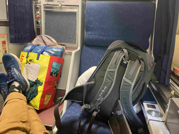 Inside the “roomette” facing a large seat, to one side is the toilet. A large backpack is on the chair and a Trader Joes bag is on top of the toilet, along with my feet (in shoes!)