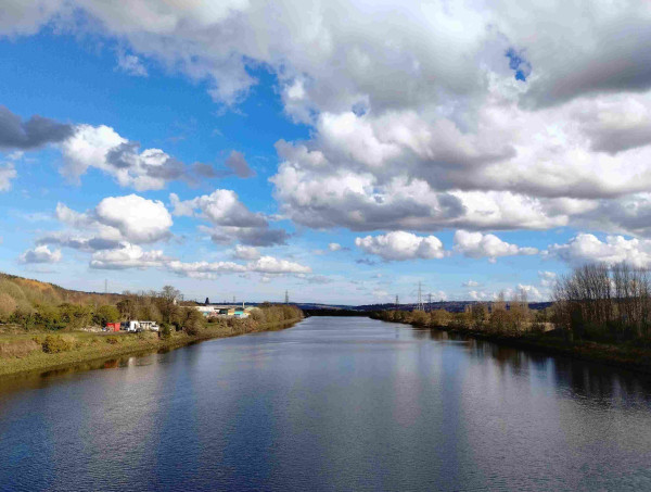 A broad river lightly rippled beneath a sky of bright blue and white fluffy clouds. The river is viewed from the middle, on a bridge (not visible). On the left bank, some small industrial units are hidden behind trees that are mostly still bare. On the right, we can see little more than trees, with a hint of farmland behind.