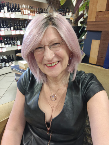 A pink haired woman sitting in a cafe wearing a black leather cap sleeve dress with a rose gold zip down the front.
Also wearing a rose gold circle pendant necklace and matching earrings.
Wine bottles are on shelves behind.