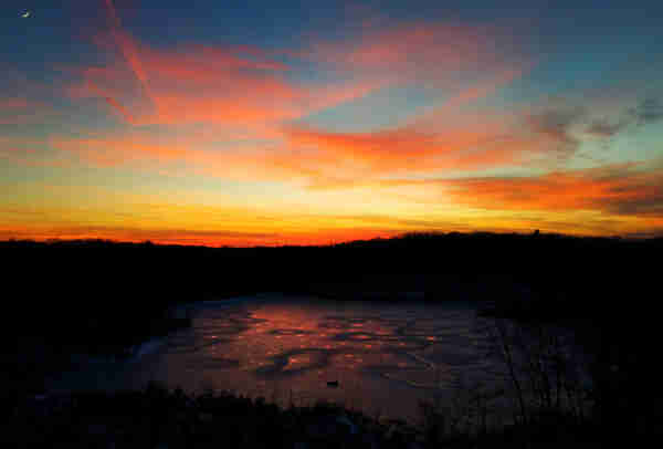 We are standing atop a ridge, looking down at a small frozen lake surrounded by low wooded ridges silhouetted in the evening light. It is past sunset, the sky is a pale blue along the horizon, deepening as you look up towards the crescent moon. A number of streaky, hazy clouds are still catching bright pinks, golds and oranges from the sun below the horizon. The surface ice on the lake is very thin and reflects darker and more muted versions of the sky colors. It has melted all the way through in many places, leaving small holes where water mirrors the brighter colors.