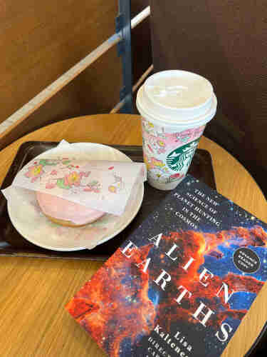 A photograph of a small table with a pink floral designed Starbucks cup and pink doughnut, wrapped in a floral sheet of paper. A book entitled “Alien Earths” is on the table with a stamp saying “advance reader copy” so you can all be jealous. 
