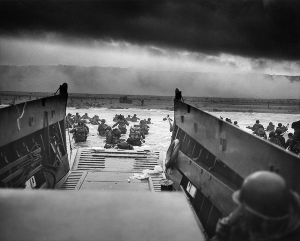 In a black-and-white photo, troops wade ashore at D-Day, observed from within the now-emptied landing craft. Beyond the open ramp at the front of the landing craft, soldiers laden with heavy packs wade to shore, their rifles held high and their backs to the camera. Perhaps 80 yards from the landing craft, the sandy beach awaits them, cluttered with obstacles, and beyond the beach cliffs covered with fog or smoke, and above the cliffs a streak of lighter sky and then a wider, ominous streak of black sky or clouds or smoke. The photo, by Robert F. Sargent, is titled “Into the Jaws of Death.”