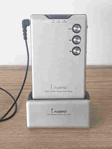 Photo of a MP3 Player (Digital Audio Player) iAudio M3 from 2004 in a cradle on a desk. A silver, metallic device, about the size of a deck of playing cards or slightly bigger than a credit card with 3 round buttons for play, forward, previous - very minimalistic, no display.