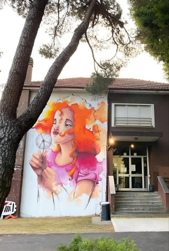 Streetartwall. The enchanting mural of a girl with a dandelion was sprayed on the outside wall of a two-storey town hall. A red-haired girl in a pink dress holding a white dandelion in her hands is depicted like a watercolor painting with flowing colors.