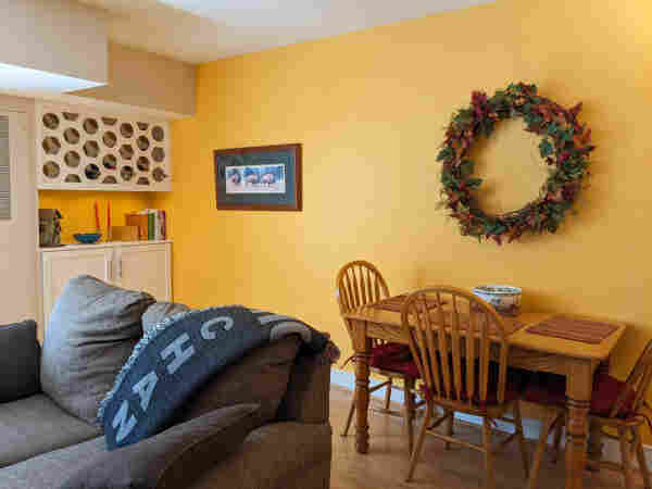 A shot of the yellow wall in our living room. There's a print of three bison and a large wreath on the wall. An oak table with three chairs sits against the yellow wall. At left is a white painted built in wine rack and cabinet with some cookbooks, a turquoise bowl and red candlesticks on top.
