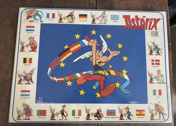 Ein altes Poster. Darauf hält Asterix eine Schriftrolle, in der alle Flaggen der EU von 1991 zu sehen sind. Der Hintergrund ist als eine EU-Flagge mit Sternen gezeichnet. Am Rand die verschiedenen Völker aus der Asterix-Serie.