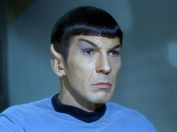 Leonard Nimoy as Spock in the original Star Trek series. He is wearing a blue shirt with a black collar, and his eyebrows are raised.