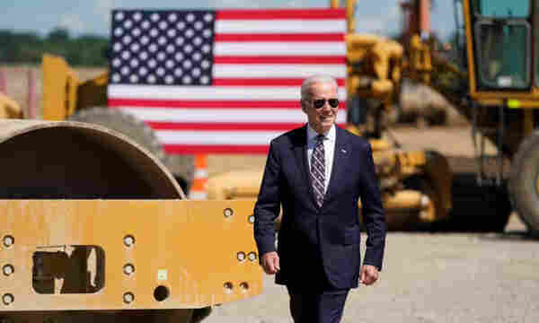 Joe Biden in front of an American flag and large machinery 