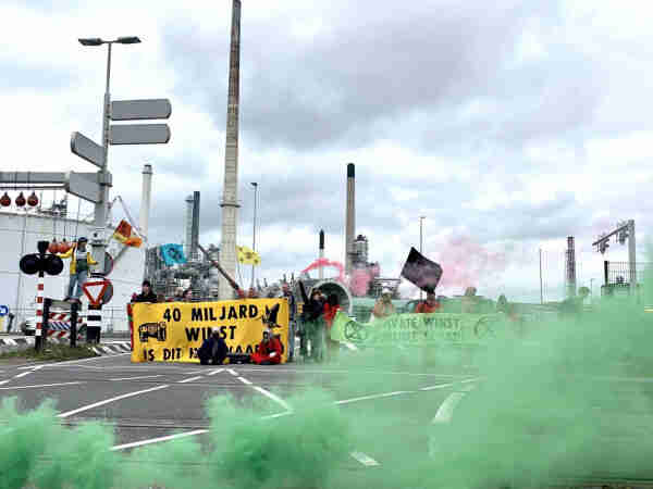 Protesters are blocking the road at Shell Pernis plant in Rotterdam. A group of people is holding signs, with green smoke blowing in the front.