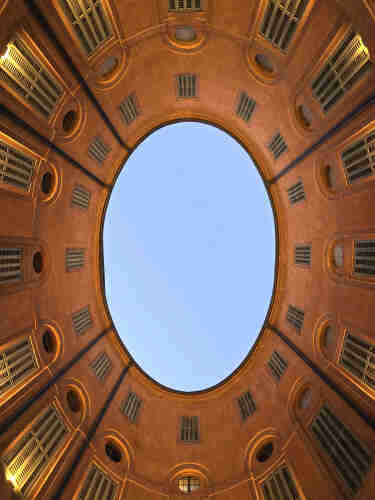 The striking view of the sky through the circular opening of a rotunda. The architecture features a series of symmetrical windows and grates, arranged in concentric circles that draw the eye upward. The warm, terracotta-colored walls glow under the soft light of the setting or rising sun, enhancing the textures and dimensions of the structure. This photograph, taken from the courtyard of the Rotonda Foschini in Ferrara, Italy, showcases the beauty of Renaissance architecture, inviting the viewer to admire the blend of functionality and aesthetic that characterizes this historic period.