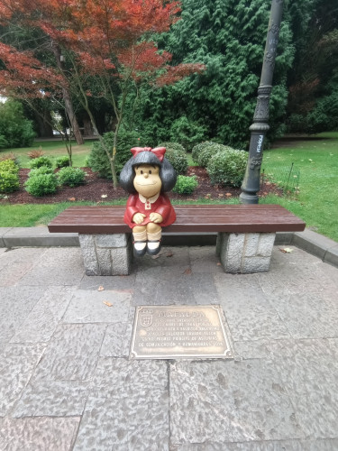 My photo of a statue of a cartoon style girl sitting on a bench. She is famous from the comics and looks like she's waiting for you to sit next to her.
Behind is a beautiful tree and a lamppost that gives the photo more depth
