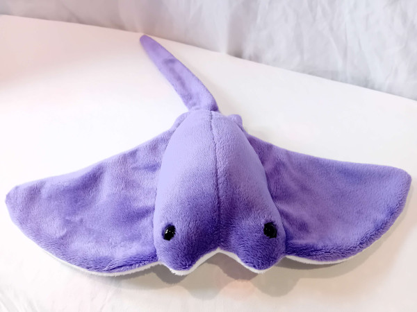 A purple plushie Seaflap (stingray) with a white underbelly, long tail and beady black eyes looking up from the top of their fishy head.