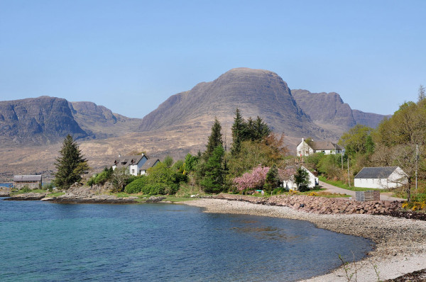 Kishorn in Wester Ross. The image shows a view over a bay with a stony beach that faces left out to sea. There’s a headland beyond the bay on which are trees and houses. In the background is a glimpse of more water, and then in the distance there are mountains with dark rocky faces rising to considerable height. The sky is blue.