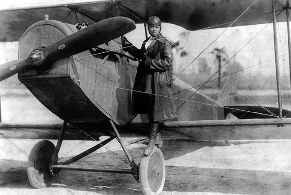 Bessie Coleman standing on the wheels of her plane in 1922. She is a black woman dressed in pilot leathers.
