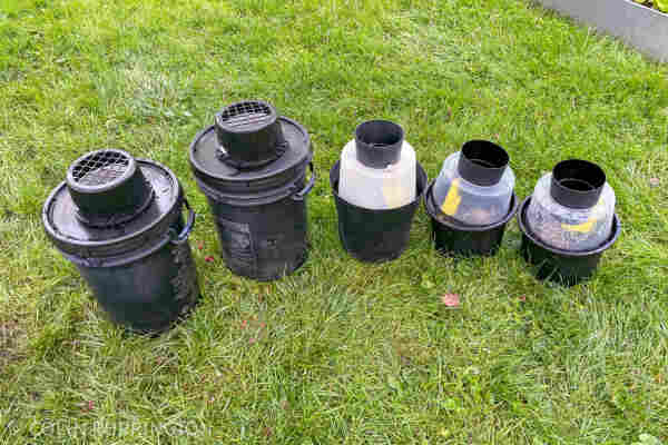 Photograph of five black buckets with various funnel-like openings through which pregnant mosquitoes can enter, lured by the stagnant water within.