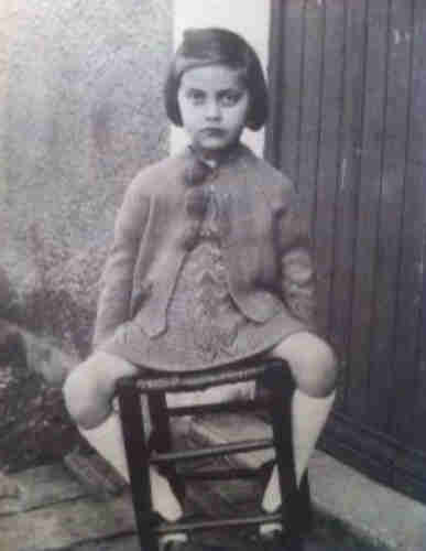 The girl sits inside on a stool. She is wearing a dress and jumper with pompom embellishments. On her feet she is wearing knee socks. Her dark hair reaches behind her ears. Her arms are placed along her body. Her hands are hidden behind her thighs. She has a neutral expression on her face. She looks straight into the camera.