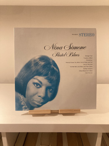 A vinyl record cover for Nina Simone's album "Pastel Blues" displayed on a wooden stand. The cover features a blue-toned portrait of Nina Simone and a list of songs.