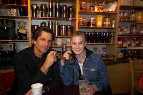The actors known for their different depictions of Starbuck (Dirk Benedict, from the original late 70’s/early 80’s Battlestar Galactica TV show & movie, and Katee Sackhoff from the ‘oughts cable/streaming series), both holding signature cigars both of their iterations of the character would enjoy, sitting at a table in a retail Starbucks location, with drinks from said chain in front of them. They are each grinning knowingly into the camera, probably wondering how long it will be until they’re asked to put the cigars away.