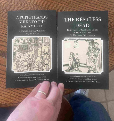 Two books with illustrated covers titled "A Puppet Hand’s Guide to The Rainy City" and "The Restless Dead," displayed on a wooden surface with a person's hand resting on the left book.