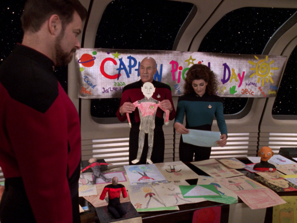 Jean-Luc Picard, Deanna Troi und Will Riker stehen in einem Aufenthaltsraum der Enterprise vor einem Tisch voller Figuren und Bildern, die Captain Picard zeigen. Im Hintergrund hängt ein Banner auf dem "Captain Picard Day" in kindlicher Schrift steht.