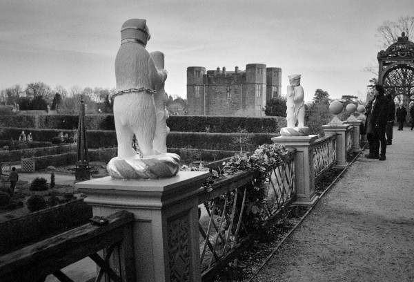 Bottom right is a terrace with a wooden balustrade. Two white chained bears stand on plinths in the balustrade, each holding a trimmed piece of wood; together they were the symbol of Robert Dudley, who owned the Castle and wanted to marry Elizabeth 1st. Between the bears, the Castle gatehouse can be seen. Black and white photo.