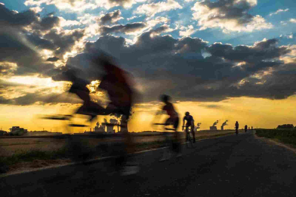 Das Foto zeigt eine auseinander gezogene Gruppe von Fahrradfahrern. Die beiden ganz vorne verschwinden fast komplett in der Unschärfe, während die hinteren Fahrer immer besser zu sehen sind. 
Im Hintergrund ist ein Kraftwerk zu erkennen, der Himmel ist bewölkt. Die Sonne scheint durch eine Wolkenlücke und taucht die Szenerie in fast goldenes Licht. 