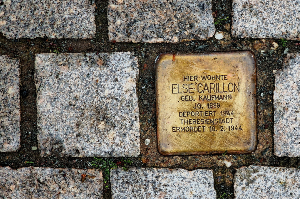 Close up shot of a pavement. One of the stones is replaced by a golden plaque, a so called 'Stolperstein', a 'stumbling stone', remembering a Jewish citizen of Nazi-Germany, who lived there before being deported to a concentration camp. 
The text reads: "Hier wohnte Else Carillon, geb. Kaufmann, JG. 1889, deportiert 1944, Theresienstadt, ermordet 16.2.1944".
(translation: "Here lived Else Carillon, nee Kaufmann, born 1889, deported 1944, Theresienstadt, murdered 16.2.1944)