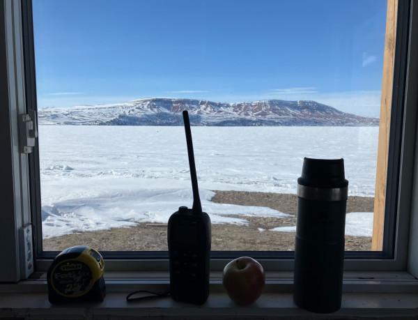 A measuring tape, VHF radio, Apple, and coffee mug sit on a window sill. In the background a frozen ocean, mountains, and blue sky. 