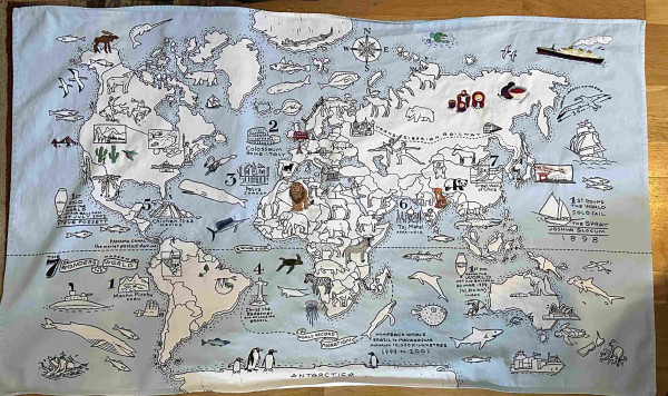 A large map of the world printed on fabric. There are lots of animals and landmarks on it. Some of them have been coloured in with embroidery. 