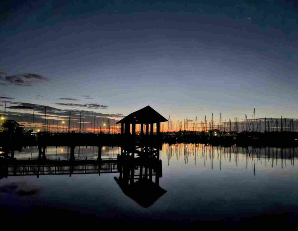 View over still, dark waters with silhouettes of sailboats and a marina with colorful lights before orange and yellow bands rising from the horizon, all casting reflections upon the water below, a long wooden pier with a covered observation deck also in silhouette casts a reflection upon the water below.