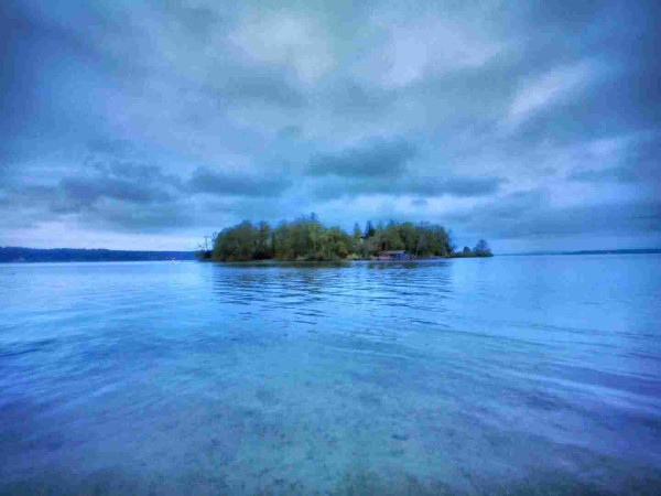A small island with trees and a structure in the middle of a calm lake under an overcast sky.