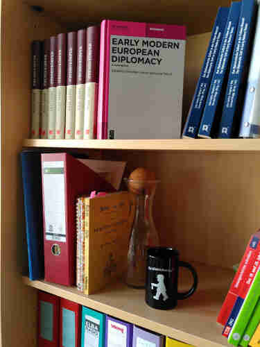 book case with several books. You see prominently the Early Modern European Diplomacy handbook on the top shelve.