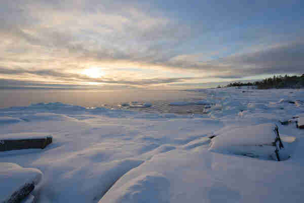 A snow covered rocky sea-shore with a frozen sea and the sun sitting very low