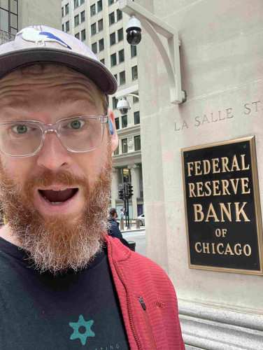 A red/grey bearded nerd with transparent glasses and a full baseball cap warning outside a sandstone building which has the plaque “federal reserve bank of Chicago” written in gold letters