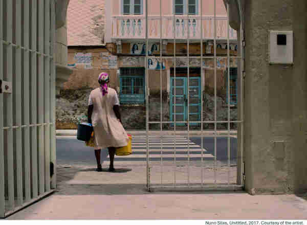 Photograph by Nuno Silas, taken in 2017. The photo shows a black woman, her back to the camera, going out of a gate carrying buckets in both hands. She is standing by a cross walk. On the other side of the street, you can see a pink building whose ground floor is very dilapidated and the first floor seems to have been restored, with a pink wooden balcony and light blue wooden doors.