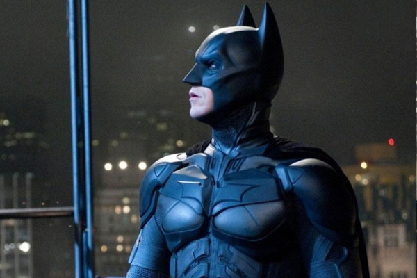 Batman, dressed in his iconic black suit and cowl, stands against a nighttime cityscape, looking out with determination.