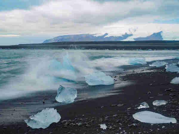 Pieces of iceberg on a black sand beach. A wave is crashing on some of them. There's a snow-topped mountain in the background. The sky is cloudy. 