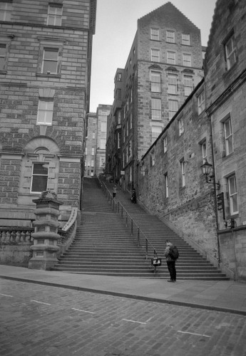 Black and white portrait format photo showing a broad set of stone steps with a metal hand-rail in the centre, rising towards a distant street between stone buildings. A man checks his phone at the bottom; other people can be seen further up. There is perspective convergence from pointing the wide angle lens slightly upwards.