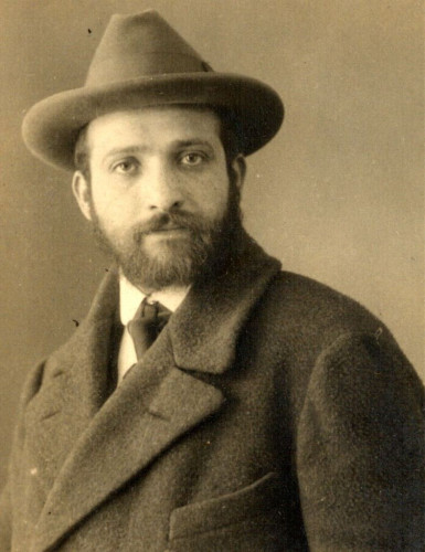 Vintage sepia-toned photograph of a bearded man wearing a fedora and a thick overcoat, positioned in a formal portrait setting.