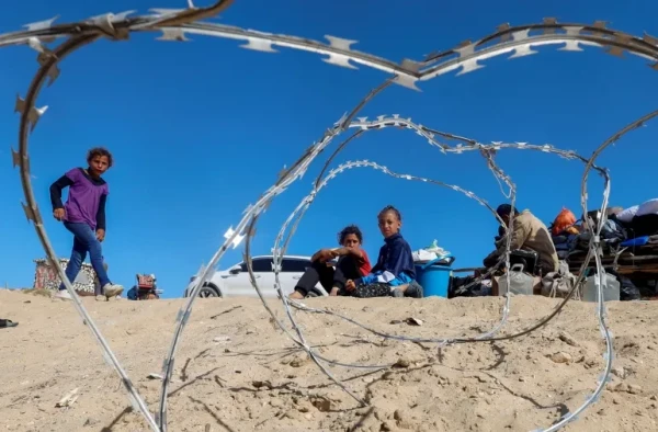 Children sitting on sand with their meagre belongings behind razor wire.