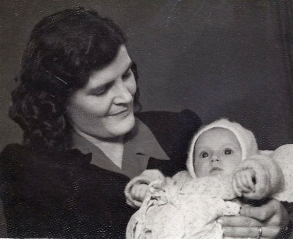 A vintage black and white photo showing a woman lovingly holding a baby wearing a knitted outfit and bonnet. The woman is smiling gently at the baby girl, who is looking directly at the camera.