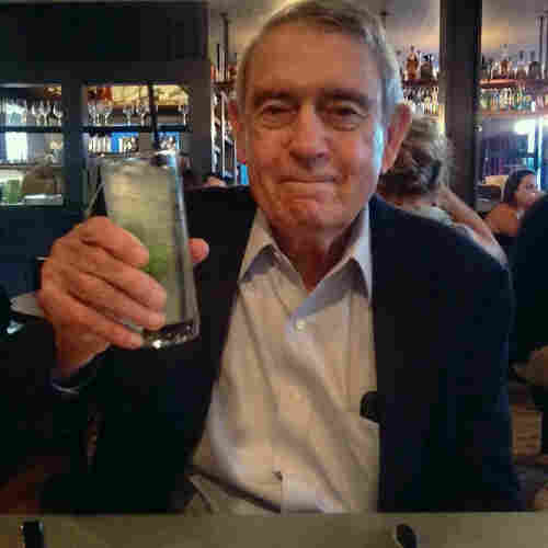 Dan sitting at a table with a bar in the background. He is smiling and holding up what he calls a ’no-jito,’ a non-alcoholic version of a mojito. He’s searing a light dress shirt with the top button open with a black sports coat over it.