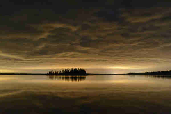 A roiling, powerfully overcast sky forms a roof over a night scene; distant city glow illuminates along the horizon and gilds the underside of the leaden sky. Below, a still, glassy lake mirrors the sky and distant forest, including a small forested island just left of centre. 